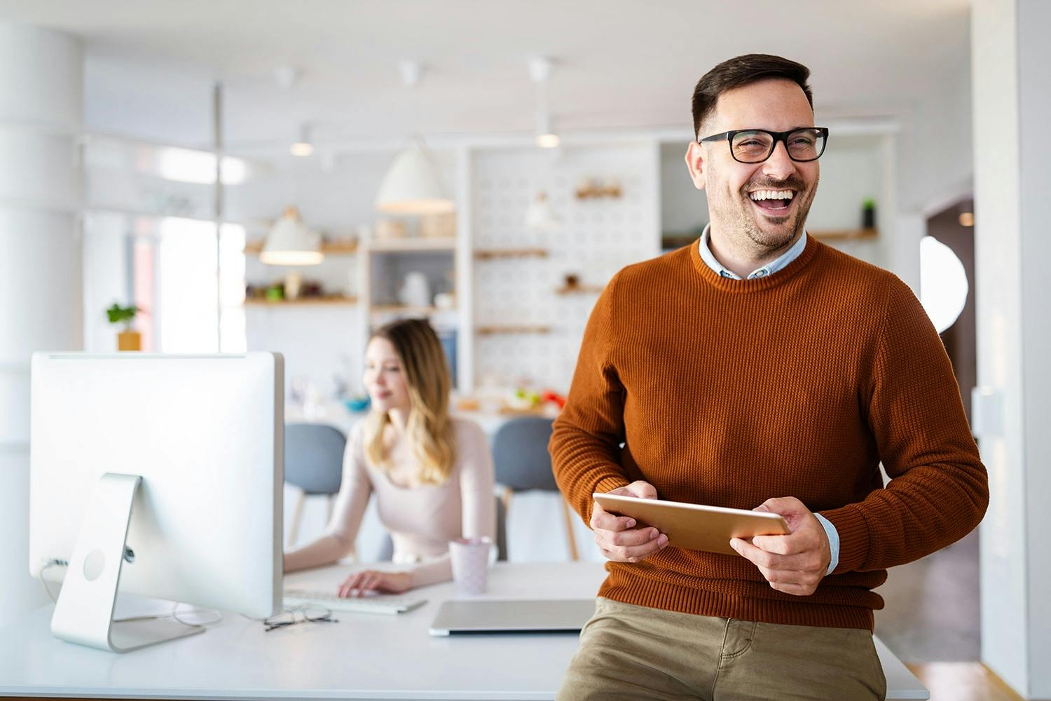 A smiling man in an orange sweater holds a tablet while standing in a modern office, with a woman working at a computer in the background.