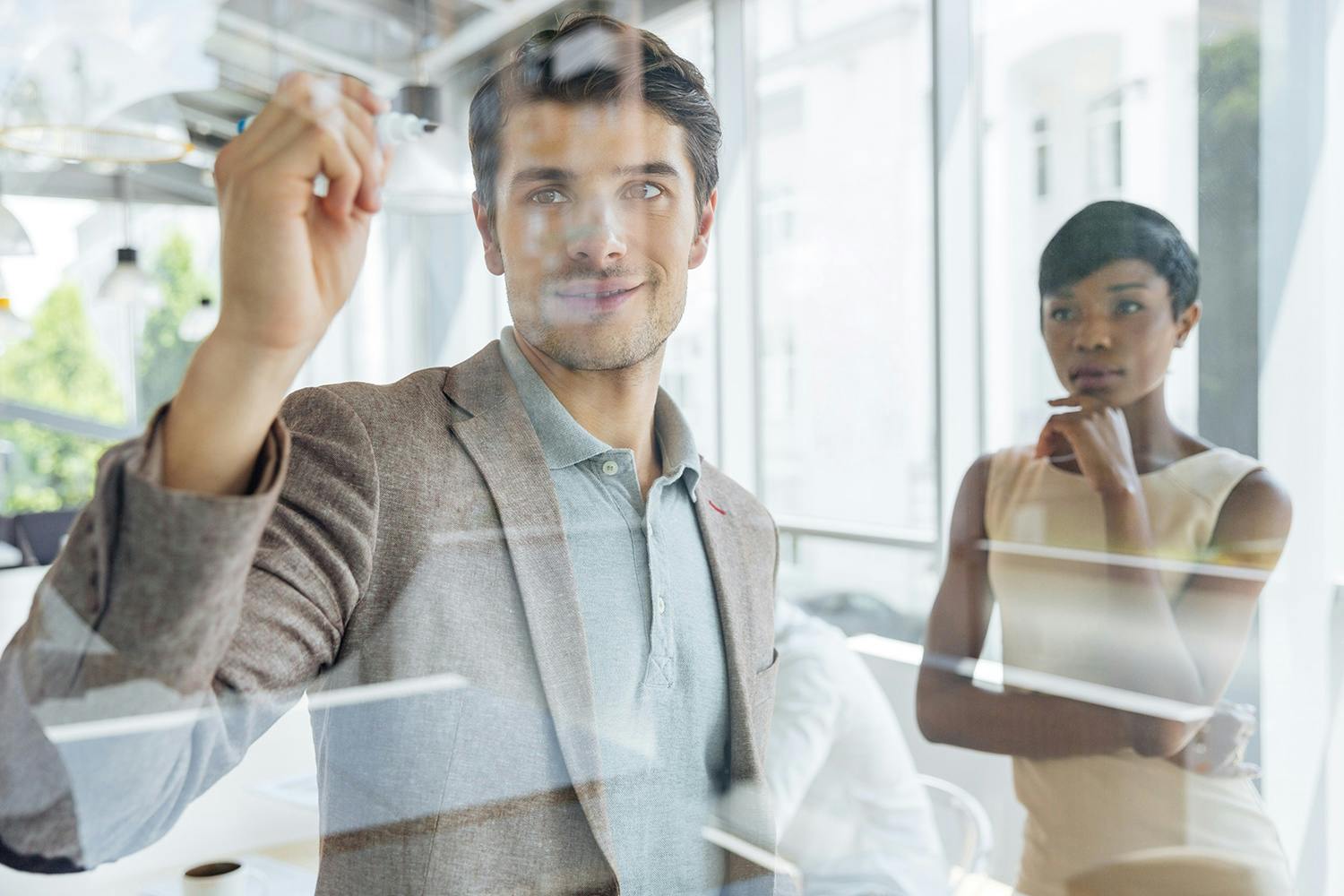 A man writes on a glass wall during a meeting, while a woman looks on thoughtfully.