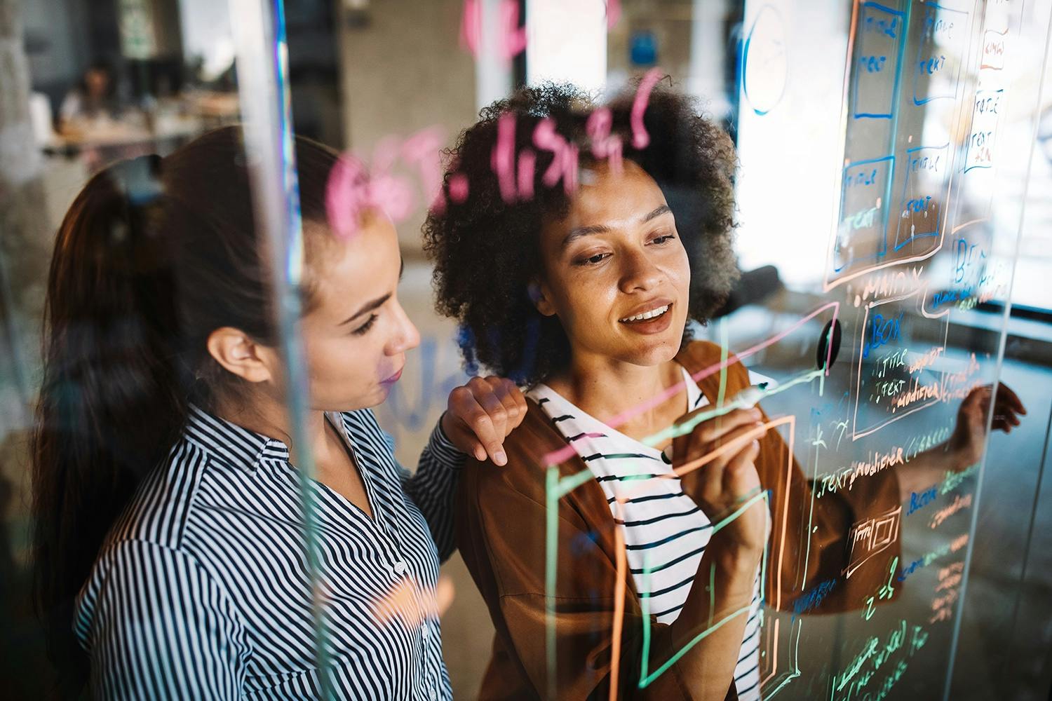 Two women collaborate on a design, writing on a glass wall with colorful markers.