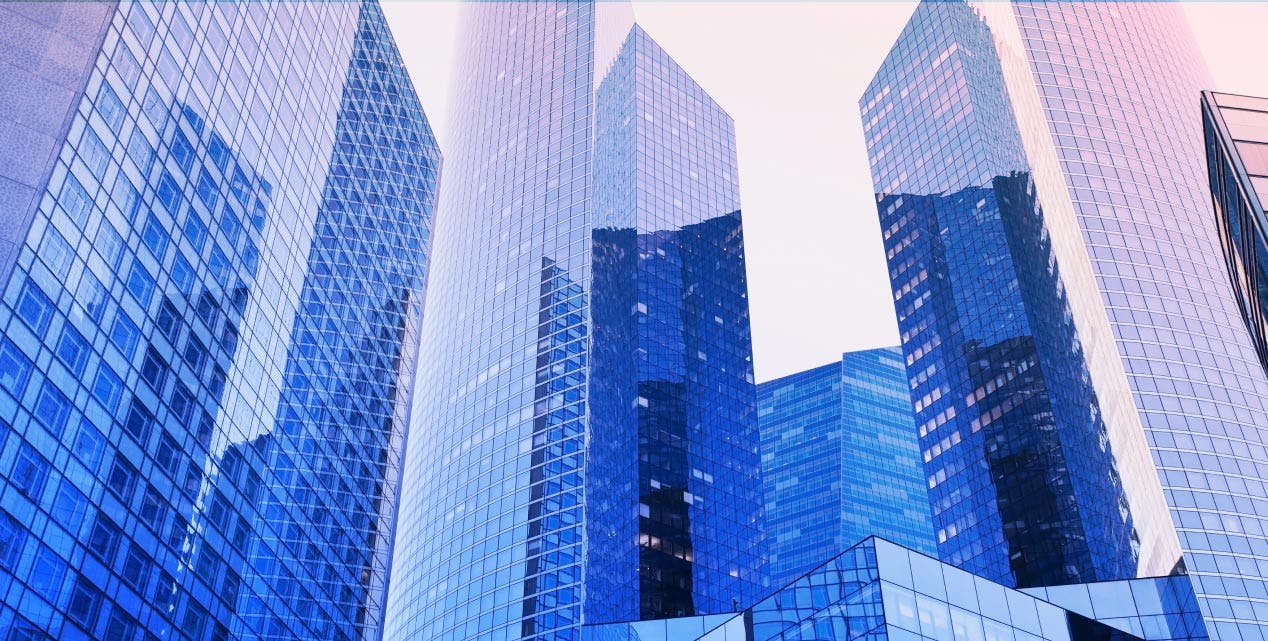 A photograph of modern glass skyscrapers reflecting the surrounding cityscape. The buildings have sleek, mirrored surfaces in shades of blue, with the sky appearing in soft pastel hues. The perspective is from below, looking upward, emphasizing the height and architectural design of the structures.