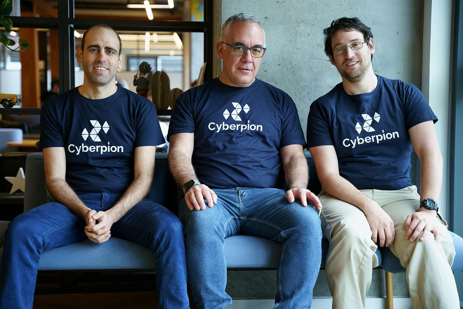 Three men sit side-by-side on a couch, each wearing a dark blue t-shirt with the Cyberpion logo. They appear to be colleagues.