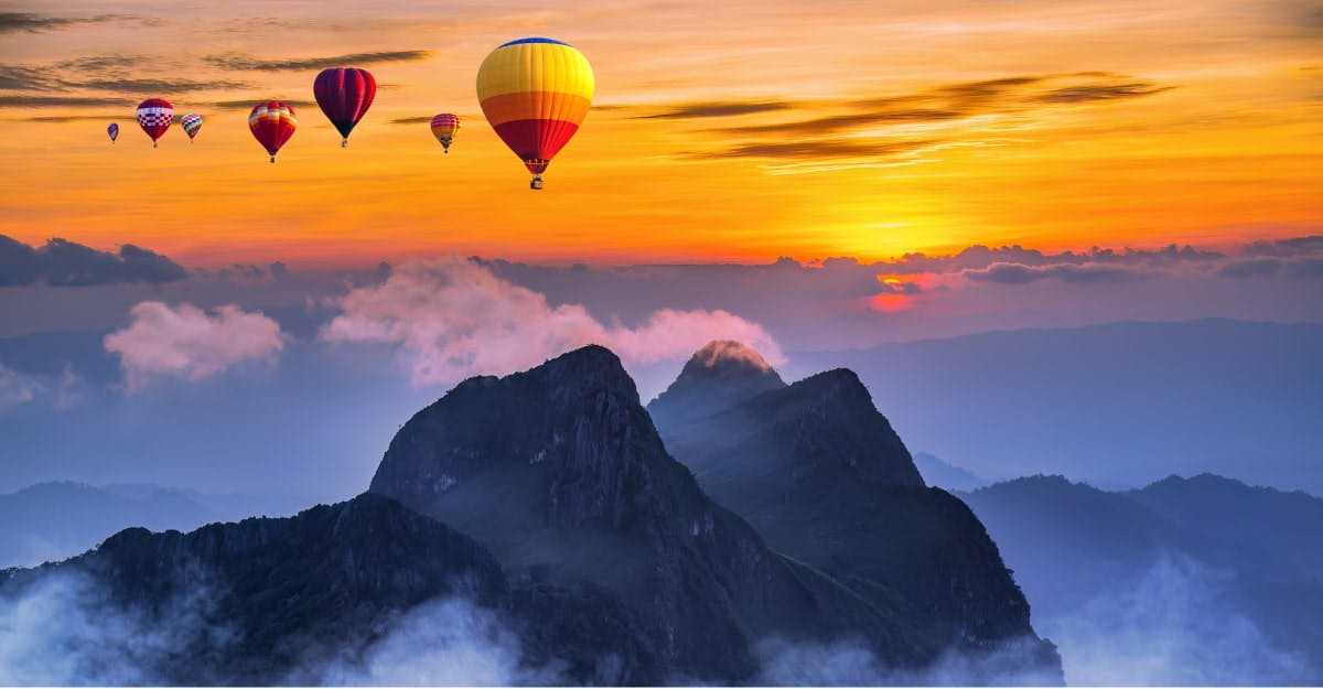 Hot air balloons fly over a mountain range at sunset.