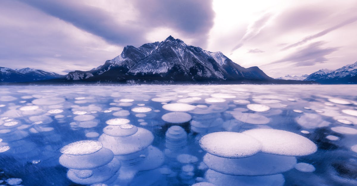 Frozen lake surface with numerous round ice bubbles, a snow-capped mountain range in the background under a cloudy sky.