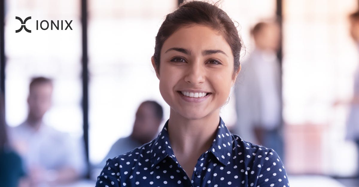 Close-up portrait of a smiling woman with dark hair, wearing a navy blue polka dot shirt, in a blurred office setting.