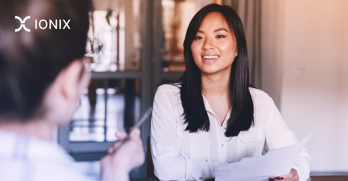A woman sits at a table, smiling and looking at someone off-camera. She is reviewing documents and appears to be in a meeting or interview.