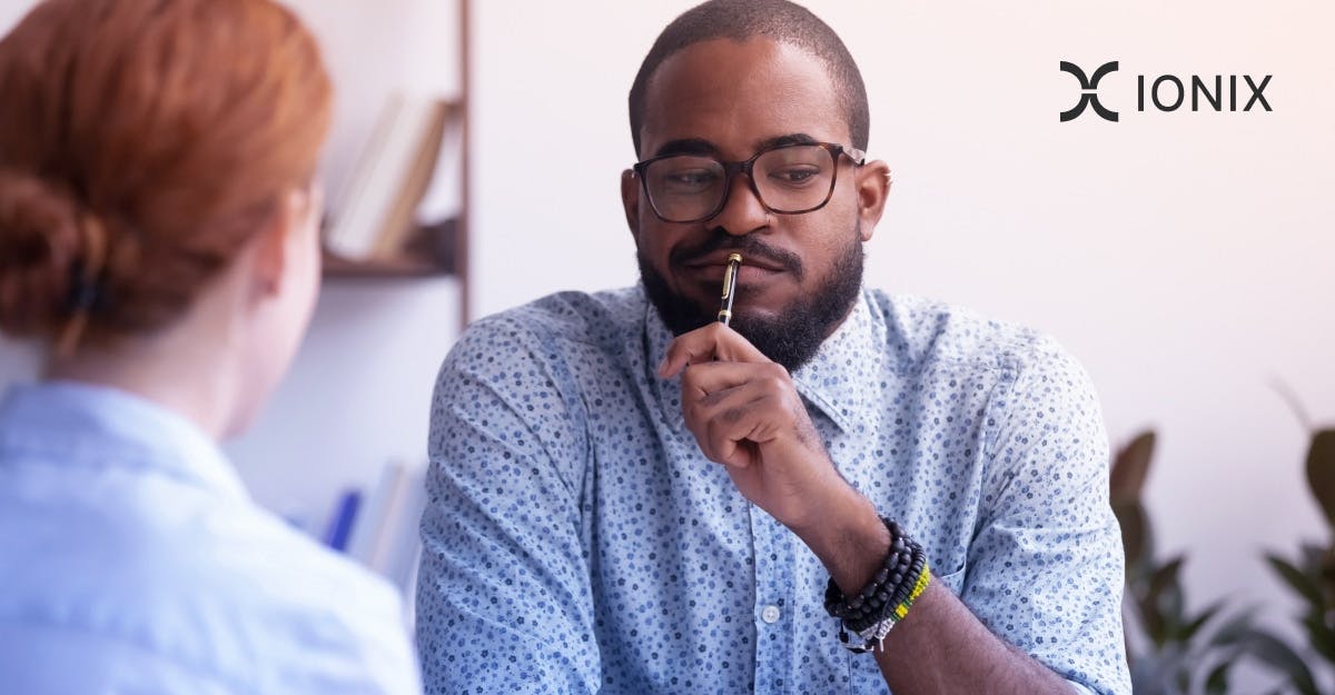 A man in glasses sits in a meeting, pensively holding a pen to his lips, listening to a woman whose back is to the camera.