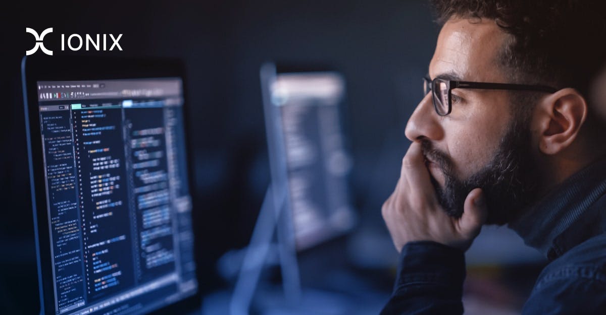 A bearded man wearing glasses sits in front of a computer screen, deep in thought as he reviews lines of code. The Ionix logo is visible in the upper left corner.