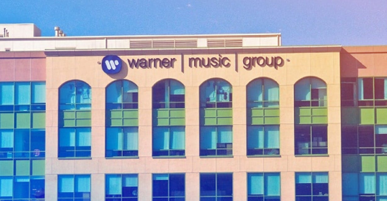 A photograph of a Warner Music Group office building with a beige facade and large arched windows. The company’s logo, featuring a blue 'W' icon, is displayed alongside the text 'warner music group' in black letters. The sky in the background is clear and blue, with a slight gradient effect applied to the image