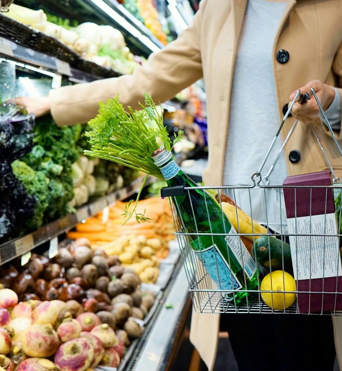A shopper in a grocery store holding a wire basket filled with fresh produce, a bottle of sparkling water, and packaged goods. The basket contains items like a bunch of celery, a lemon, an avocado, and a baguette. The person, wearing a beige coat, is reaching for leafy greens from a shelf stocked with fresh vegetables, including kale, carrots, and beets.